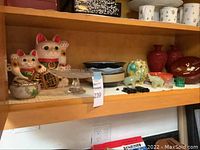 Wide shelf view showing all the Asian decorative items together: lucky cats, bowls, vases, elephants, cake stand.