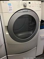 Front view of white Maytag front-load dryer mounted on matching storage pedestal, showing control panel and round glass door