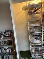 Full side view of the tall white wire mesh organizer showing its five wire baskets and contents inside. The organizer is next to a bookshelf with DVDs and CDs.