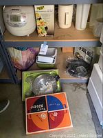 Shelf with Zojirushi rice cooker, ceramic pitcher, plastic containers, boxed salad maker, glass lid cookware, and black plastic ware