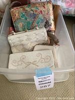Clear plastic bin containing four small handbags; floral textured multicolor bag at the back, white beaded purse, cream scallop beaded clutch, white clutch with decorative swirl pattern in front.