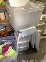 Large translucent plastic storage bin on top of stacked white plastic drawers, next to folded panels or boards.