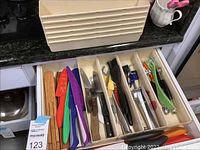 Photo showing drawer open with bamboo subdividers, colorful knives with sheath, various kitchen knives, peelers, and measuring spoons in cream plastic drawer organizers.