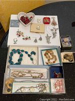 Photo of assorted costume jewelry featuring Christmas-themed earrings, bracelets, necklace sets, men's cuff links, and key chains arranged on a white surface with some items in small boxes and bags.
