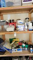 Shelf with kitchen appliances such as red blender, white juicer, silver coffee pot, and kitchen utensils; garden tools and sprayer on lower shelf.
