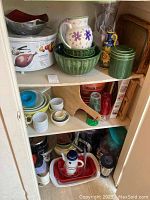Wide view of closet shelf with various kitchenware including metal mixing bowls, green ceramic mixing bowls, floral pitcher, canisters, glassware, mugs, wooden cutting board and slow cooker on top shelf.