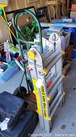Little Giant extension ladder folded and secured to green-handled hand truck, ladder shows label and locking mechanism.