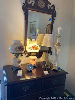 Photo showing five table lamps and Baby Ben Westclox desk clock on antique style dark wood cabinet beneath an ornate mirror.