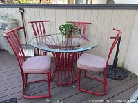 Patio set arranged on a porch deck; 4 red metal chairs with cushions and round beveled glass tabletop on red metal pedestal base.
