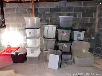Stacked plastic storage bins and totes in basement with concrete block wall background. Various shapes, sizes, and colors mostly gray, clear, and green-lidded boxes.