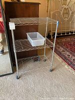 Metal wire rolling storage shelf shown from front angle with clear plastic bin on middle shelf near a wooden dresser and bed frame.