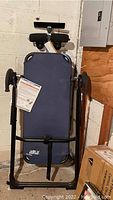 Folded Teeter Hang Ups inversion table shown upright against a wall, displaying blue padded surface and black metal frame.