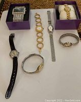 Photo taken showing 7 assorted watches with varied band styles and colors, some in purple boxes