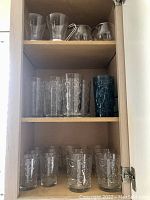 Photo showing midcentury glassware arranged on three wooden shelves inside a cabinet. Top shelf holds two glass pitchers and three clear short glasses. Middle shelf has tall glasses and one blue textured glass. Bottom shelf displays four short clear glasses.