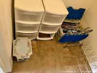 Photo showing two white plastic drawer units, blue bins containing assorted hangers, small decorative stool on carpeted floor.