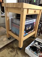 Butcher block table viewed from the side showing the thick solid wood top, single drawer, and lower shelf supporting a boxed UltraVection Oven.
