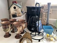 Photo showing six brown ceramic goblets, wooden birdhouse, Black & Decker coffee maker with carafe, metal kitchen stand, ceramic salt and pepper shakers, small blue ceramic bowl, wooden Happy Harvest plaque
