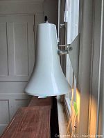 Side view of a white bell-shaped metal wall light fixture mounted on a metal bracket on a window frame, showing the shape and mounting.