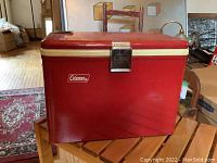 Front view of the vintage red metal Coleman cooler showing the Coleman logo on the front panel and cream trim on the lid.