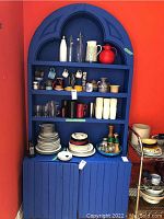 Front view of the blue painted cabinet in kitchen corner filled with various kitchenware items including plates, mugs, pitchers, and bowls.
