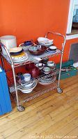 Metal utility cart with three wire shelves, holding various bowls, plates, and cups (contents not included in sale). Cart against orange wall and sitting on wood floor.