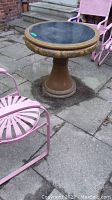 Full view of the round table outdoors, showing pink metal chairs beside it and the stone tile ground.