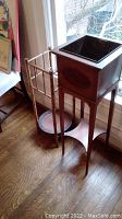 View of rattan plant stand next to antique wooden plant stand with metal insert on a hardwood floor by window.