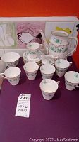 Full tea set arrangement showing coffee pot without lid, seven cups, eight saucers, creamer, and sugar bowl, all decorated with floral patterns and gold trim.
