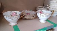 Two Royal Crown Derby teacups (white with pink floral and gold details) and six matching saucers arranged on shelf.