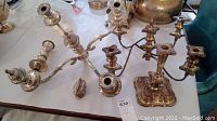 Wide shot of two large ornate silver plate candelabras, additional candle holders, and lighter on table with some silver wear.