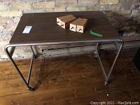 Photo showing the entire metal and laminate table with four wooden chair legs on top against a brick wall and wooden floor.