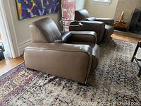 Two taupe leather chairs with black piping displayed side by side on patterned rug under wall art