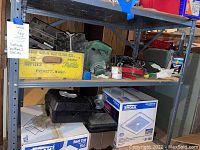 Wide shot showing metal shelving with miscellaneous tools, a yellow vintage Pepsi crate, green fan, metal brackets, and black tool box.