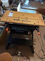 Front view of workbench showing wooden top with Black & Decker logo and Workmate branding, foldable metal legs, and orange adjustment handles.