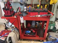 Red rolling work bench holding various tools including springs, power tool, red canvas tool organizer, and spray cans.