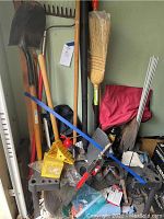 Garden tools arranged leaning against a wall including shovel, rake, broom, trimmer parts, yellow plastic holders, and assorted accessories.