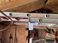 Side view of aluminum ladder with Craftsman brand label and warning labels attached, stored on ceiling hooks in a shed.
