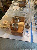 Overview of the box with pewter creamer and sugar bowl, adjacent decorative glassware, and a cream-colored floral dish.