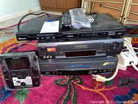 Overview of stacked video components including DVD players and VHS players with power strips on a patterned carpet.