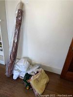 Bolt of brown fabric standing upright wrapped in clear plastic beside folded sheets and blankets on a white floor near a wall.