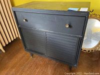 Front view of black wooden cabinet showing one drawer with round metal knobs and two slatted sliding doors.