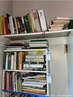 Shelves filled with various cookbooks arranged standing upright and stacked, showing a collection of diverse titles.