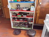 Full view of shelving unit containing various pots, pans, mixing bowls, and cookware items, with cast iron skillets and a covered cast iron pot on the floor in front.