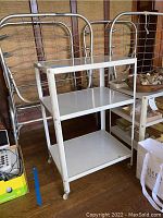 Two white metal kitchen carts with three shelves each, placed side by side, showing full profile and shelving.