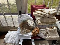Photo shows a pile of assorted linens, several lace crochet pieces, two red throw pillows, and a stack of folded blankets or linens on a chair by a door.