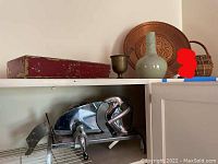 Manual meat slicer on wire shelf below a cabinet, showing shiny metal and crank handle