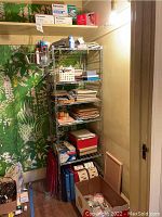 Metal shelving unit with assorted office supplies arranged on shelves in a small room with green leafy wallpaper.