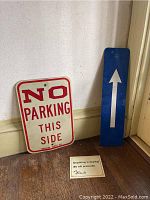 Two metal street signs ('No Parking This Side' and blue arrow) and small shoplifting sign shown together leaning against wall.