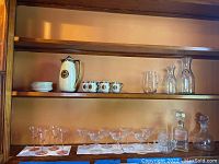 Overall display of entire lot on shelves showing porcelain tea set, clear glass pitchers, pink stemmed glasses, crystal champagne glasses, and two decanters