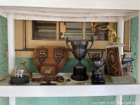 Shelved display showing multiple trophies and plaques including wooden curling championship plaque, large cup trophies, figurine trophies, and other items.
